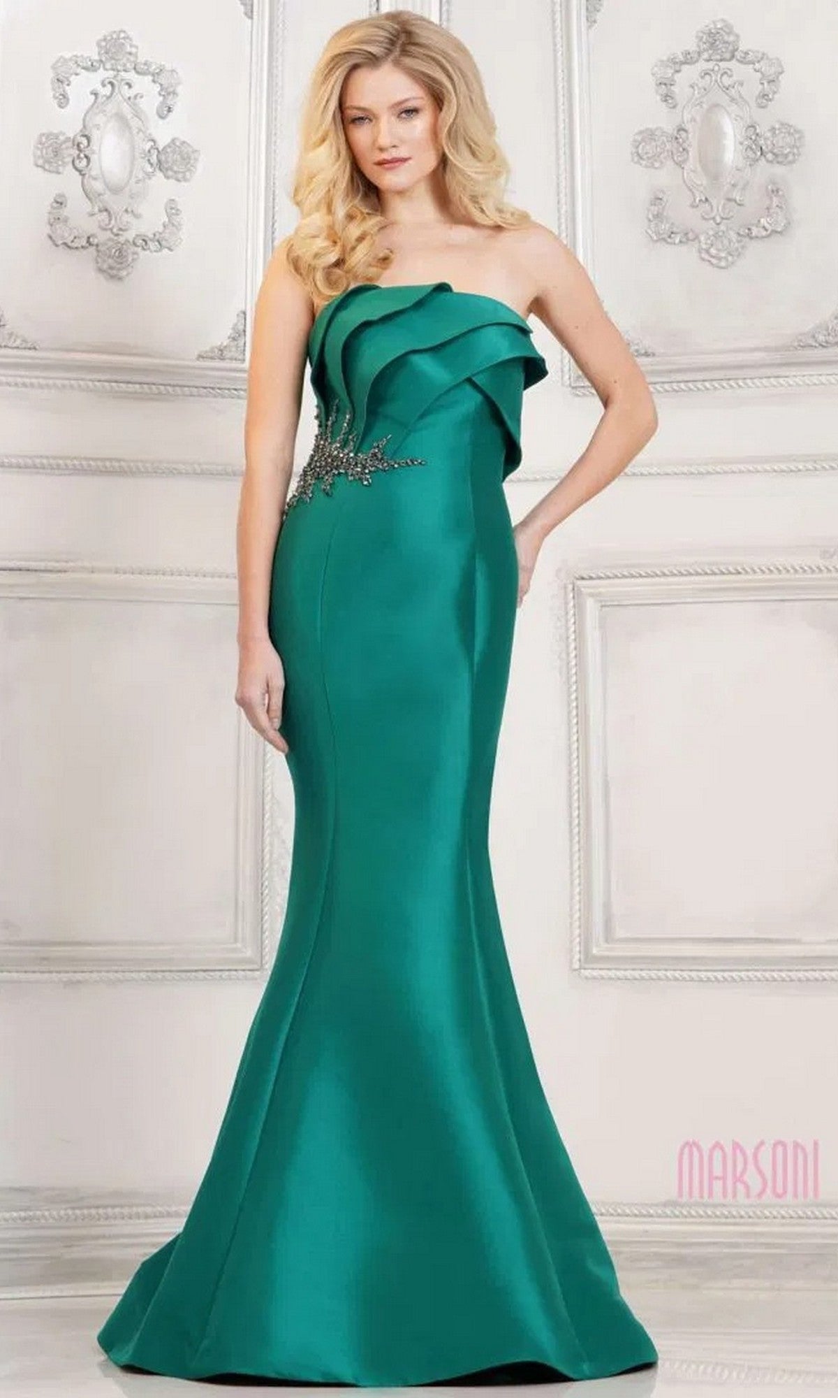 A woman wearing the Marsoni MV1308 long formal dress in emerald, featuring a strapless mermaid silhouette and ruffle details, stands indoors against an ornate white wall.
